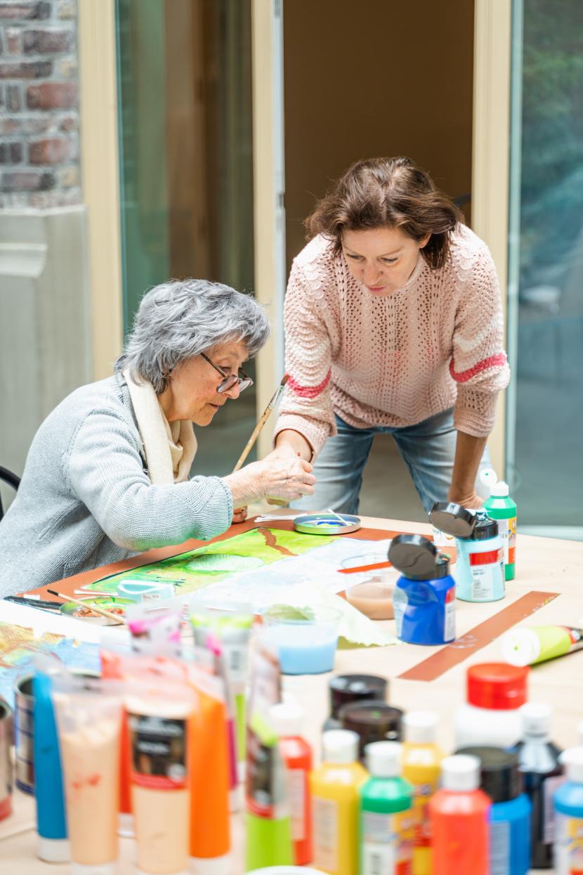 Katrien hurkt neer bij een slechtziende vrouw aan een tafel vol verftubes. De slechtziende vrouw heeft een penseel in haar hand en rond haar papier ligt een kartonnen boord zodat ze kan voelen waar de randen van haar blad zijn.