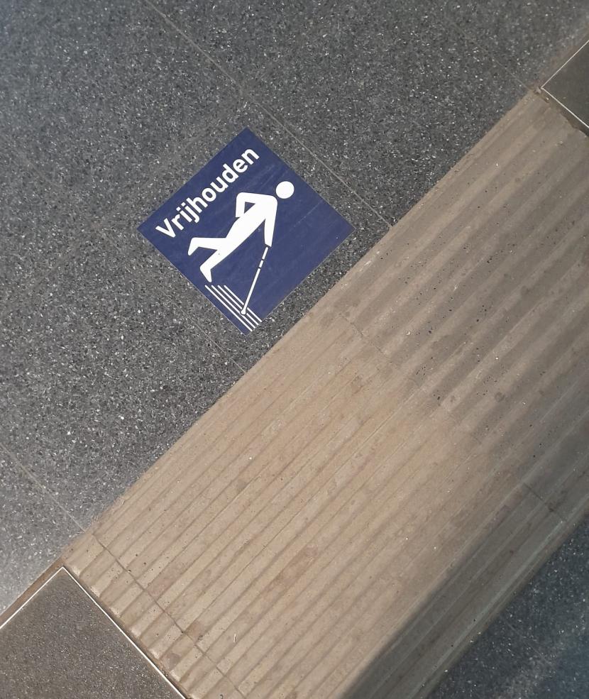 Een foto van de ribbellijnen in het station van Brugge. Ernaast kleeft een sticker met het woord 'Vrijhouden' en een pictogram van een wittestokgebruiker.