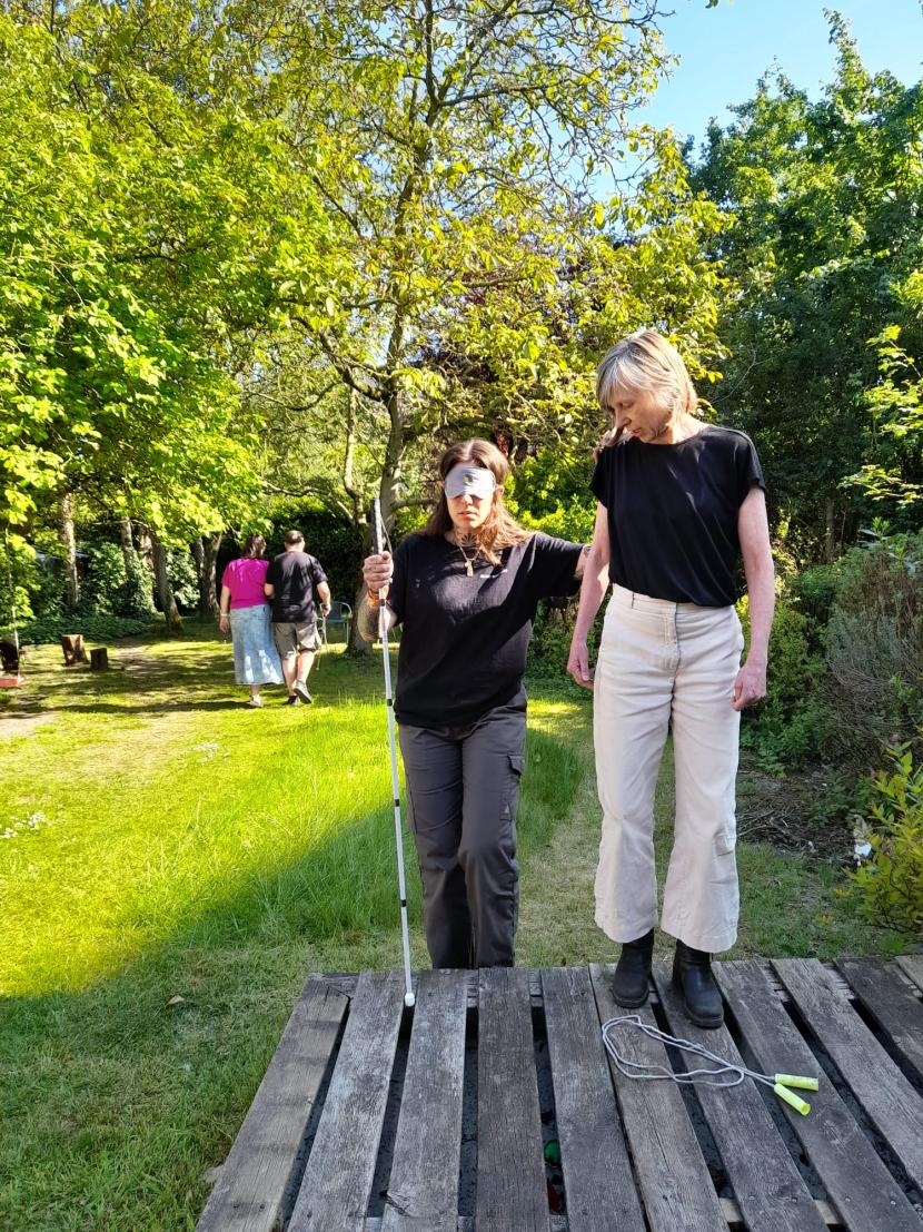 In een zonnige tuin lopen twee duo’s, waarvan telkens één persoon geblinddoekt is en een witte stok gebruikt. De tweede persoon begeleidt de andere.