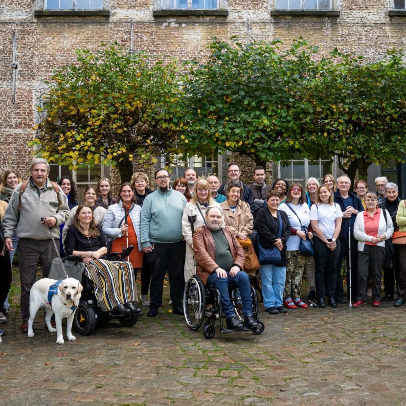 Een groepsfoto van veertig mensen in de binnentuin van de Abdij van Drongen. Er zijn twee rolstoelgebruikers en enkelen met een witte stok, er zijn ook twee geleidehonden.