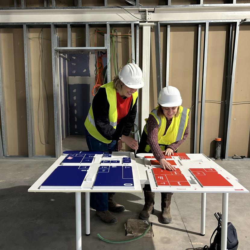 Paula van Kanal en Jo van Symfoon staan op een werf met fluohesjes aan en helmen op. Ze staan voor een voelbare plattegrond van de werf. Jo voelt aan de plattegrond.