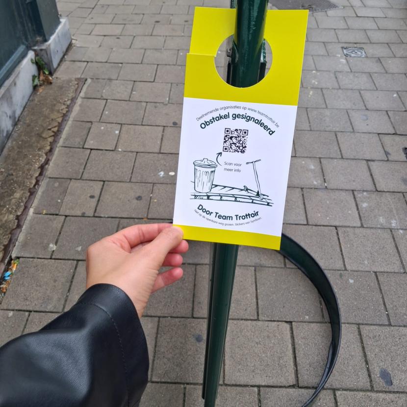 Een hanger van Team Trottoir hangt aan een promobord van een restaurant dat midden op de stoep staat.